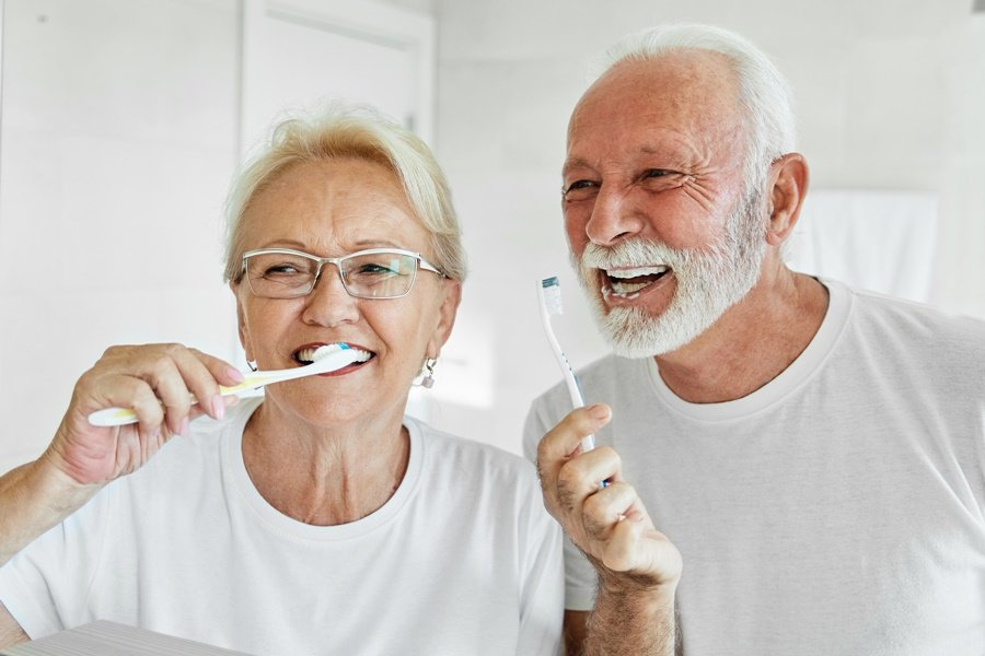 Pareja de adultos mayores cepillándose los dientes en el baño, como parte de una rutina diaria de higiene oral para prevenir el envejecimiento dental y conservar la salud bucodental.