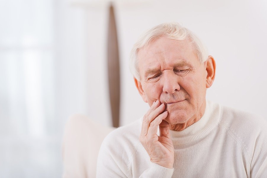 Hombre mayor con gesto de dolor mientras se toca la mandíbula, mostrando una posible molestia causada por el envejecimiento dental no tratado, como la sensibilidad o una infección periodontal.