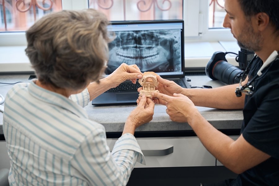 Odontólogo mostrando a una paciente mayor un modelo dental mientras observan una radiografía en el ordenador, explicando qué es un implante cigomático y cómo se coloca.