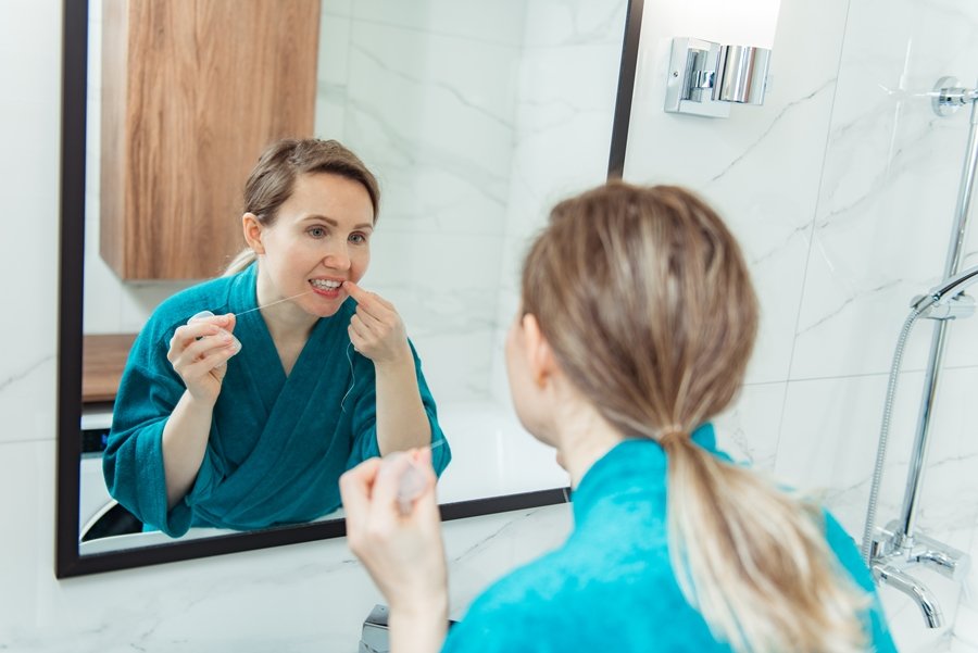 Mujer utilizando hilo dental frente al espejo del baño como parte de la rutina diaria para prevenir la retracción gingival y mantener encías sanas.