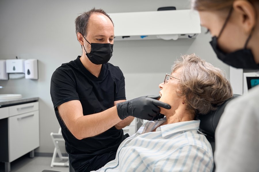Dentista examinando la mandíbula de una mujer mayor en clínica, ejemplo de revisión odontológica en pacientes con artritis y salud dental comprometida.