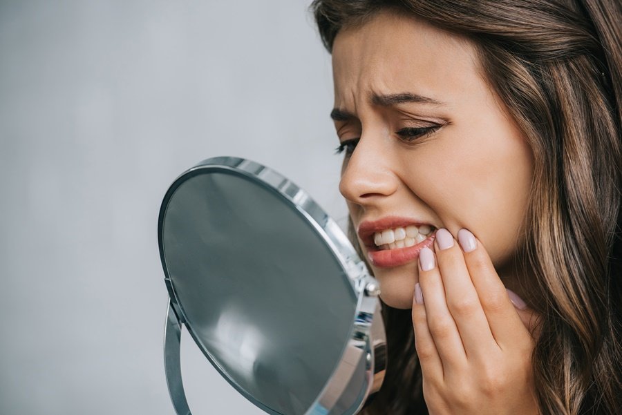 Mujer con expresión de dolor observando sus dientes en el espejo, una posible señal de caries o sensibilidad provocada por el déficit de flúor en el esmalte dental.