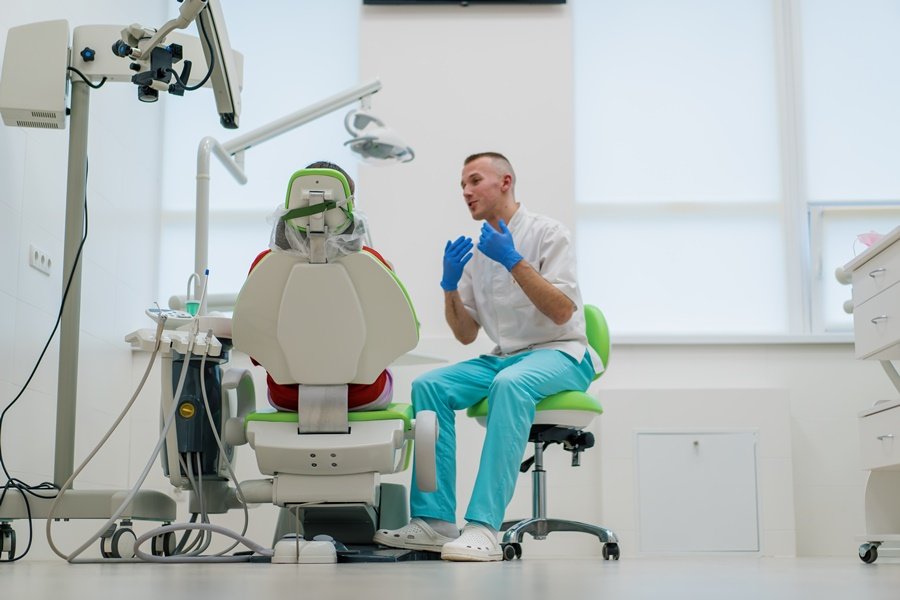 Dentista con guantes azules conversa con un paciente en consulta, en un entorno clínico moderno, subrayando la relación entre enfermedades cardiovasculares y salud bucodental.