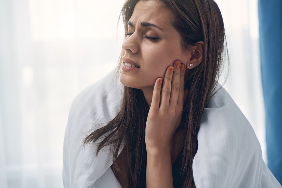 Mujer joven sentada con gesto de dolor mientras se toca la mandíbula, representando los efectos del estrés y aftas bucales en la salud oral.